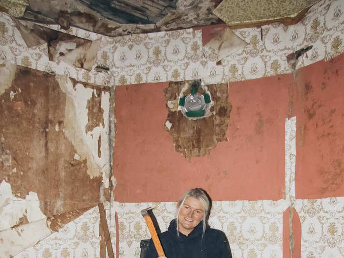 Katie posing with a hammer in a room with wallpaper peeling off