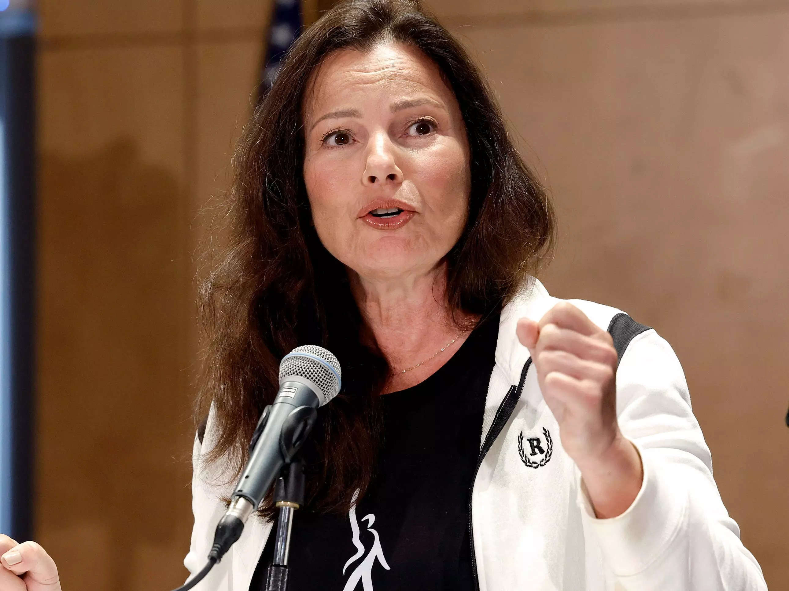Fran Drescher speaks at a microphone on a podium, wearing a white jacket, with her fist raised.