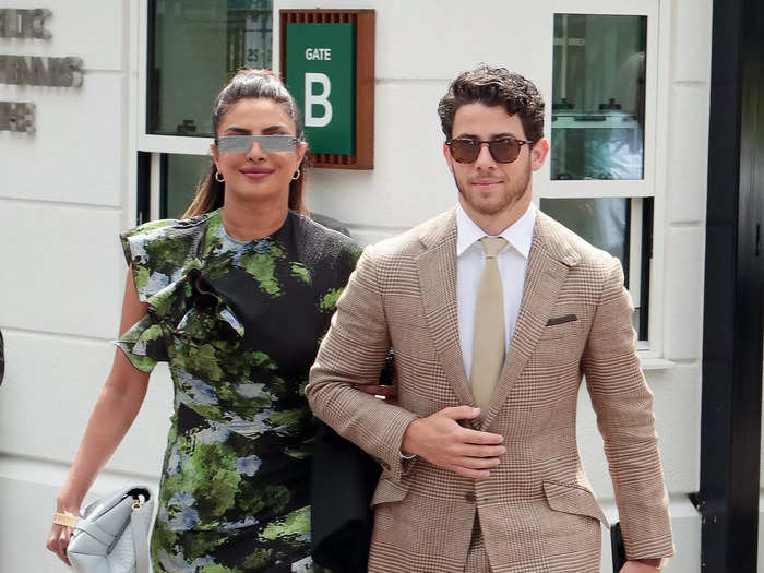 Priyanka Chopra Jonas made a statement in a black, green, and gray dress with a watercolor print and asymmetrical hemline with knee-high, open-toed, black boots and sunglasses. At her side, Nick Jonas opted for a beige, checkered suit.