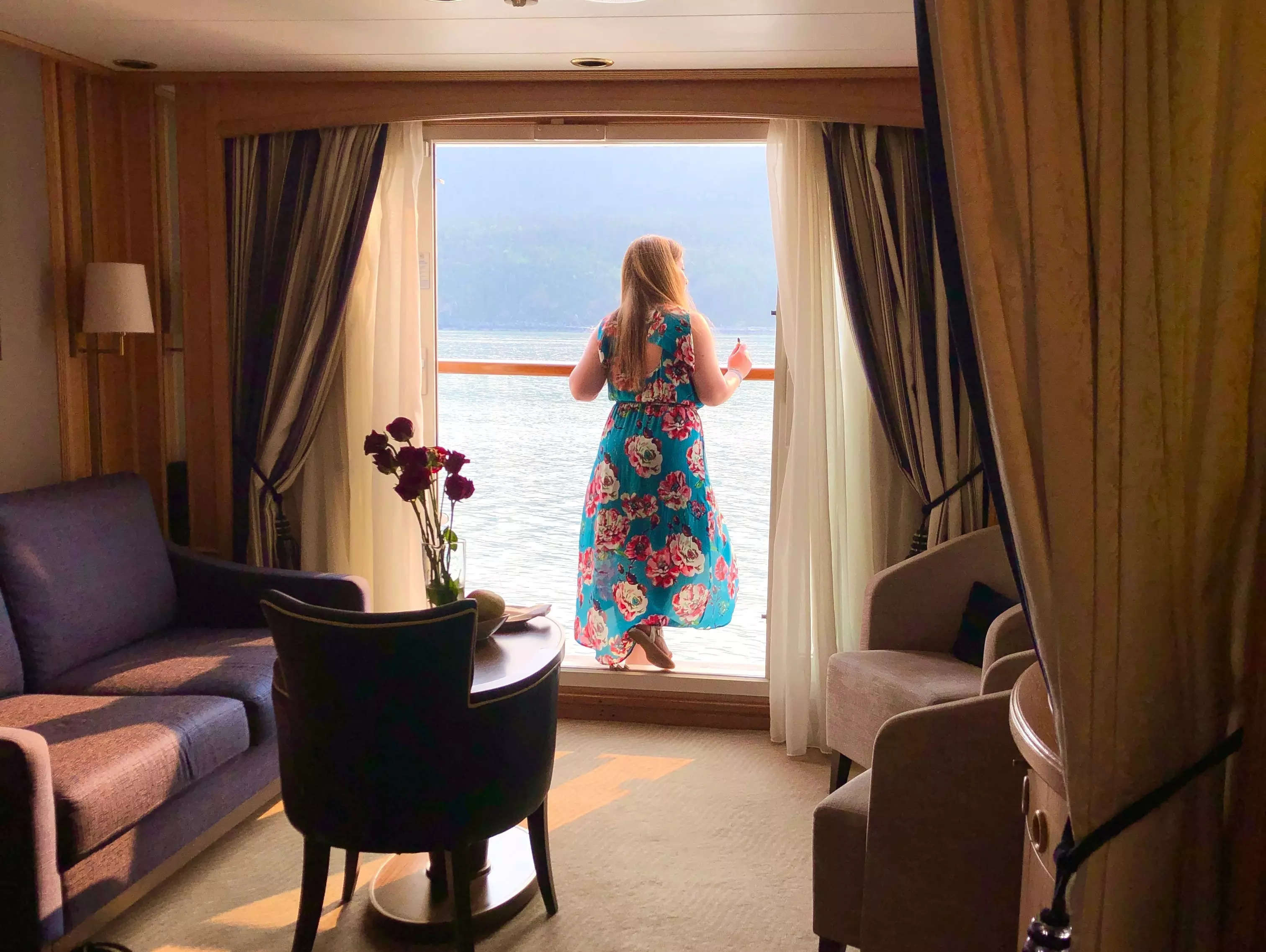 woman in floral dress standing on cruise balcony with dimly lit living room of cruise cabin behind her