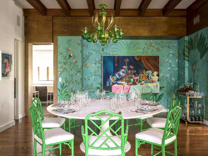 A whimsical dining room has green and white chairs, a round table, a chandelier, floral wallpaper, and a large painting.