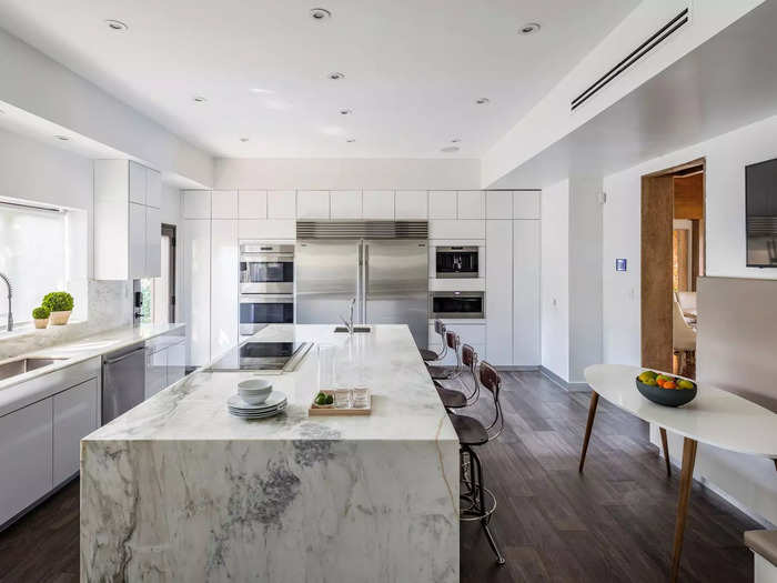 The kitchen has a neutral palette, marble counters, two sinks, a side table, and ample seating.