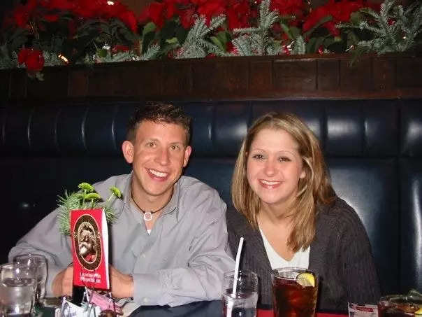 Terri Peters and her husband on their first date, sitting in a booth with navy leather seats at a restaurant. Terri has dirty blonde hair, brown eyes and wears a grey cardigan with a white t-shirt. Her husband has short brown hair, hazel eyes and wears a long sleeved grey button-down and a necklace with a black string and a white pendant.