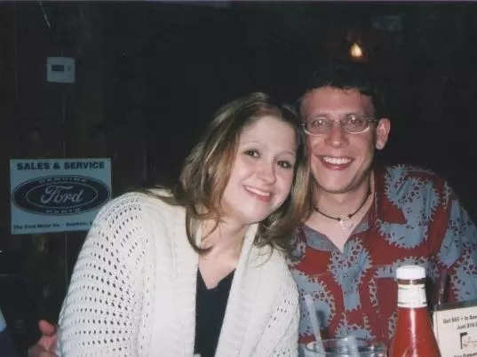 Terri Peters on the left and her husband on her right smiling while sitting at a restaurant when they first started dating. Terri wears a white knit sweater and black shirt. She leans towards her husband and he has his arm around her. Her husband wears glasses, a red Hawaiian shirt with a silver-blue abstract pattern, and a necklace with a black thread and white pendant.