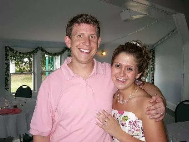 Terri Peters and her husband on her wedding day. Terri has her hair pulled back into a bun and wears a pearl necklace, pearl drop earrings, and a strapless white dress with pink roses on it. She has her hand on her husband