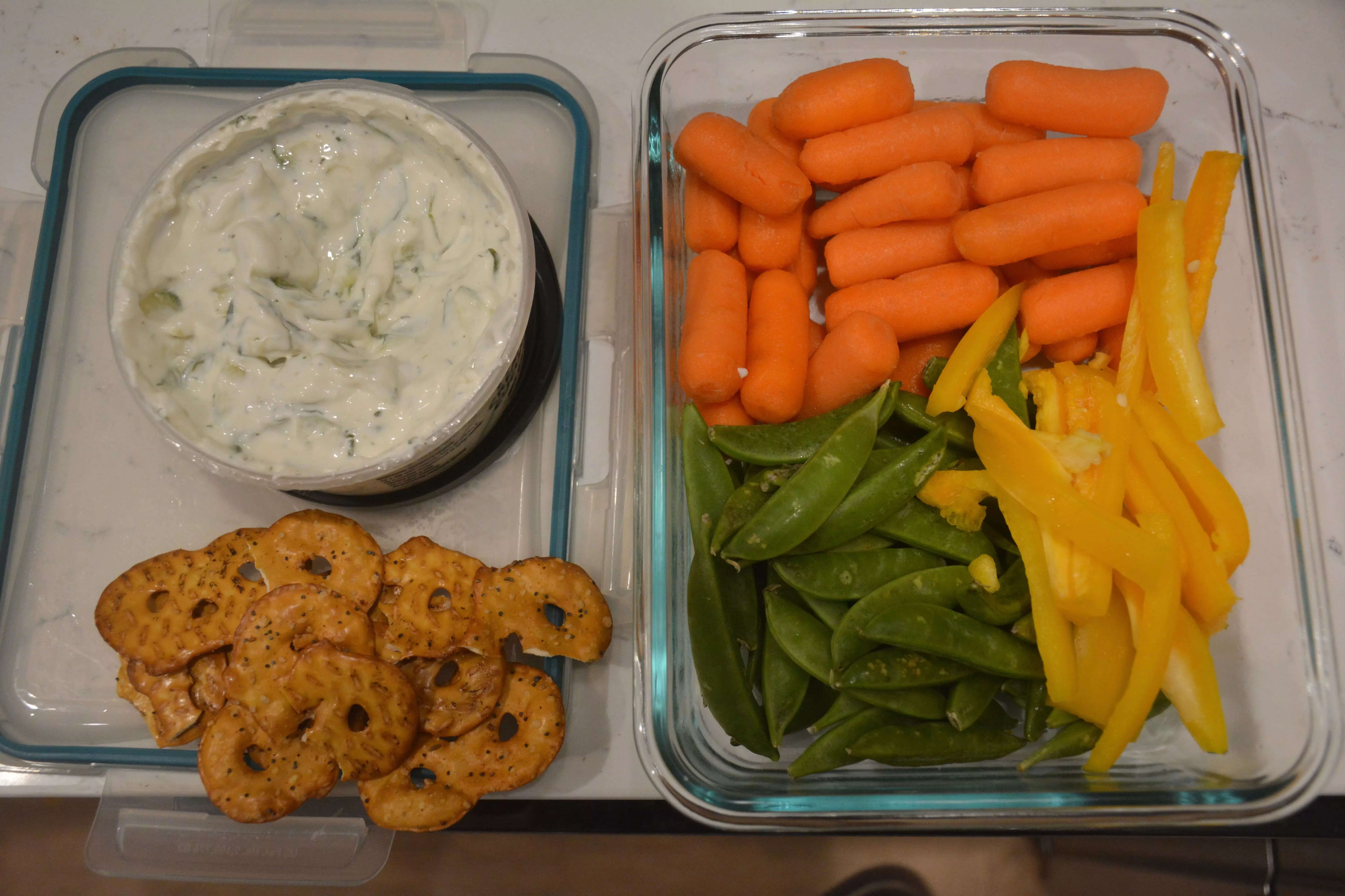 Tupperware of ready-to-eat veggies, including carrots, snap peas and yellow bell pepper, next to tzatziki and pretzel crisps.