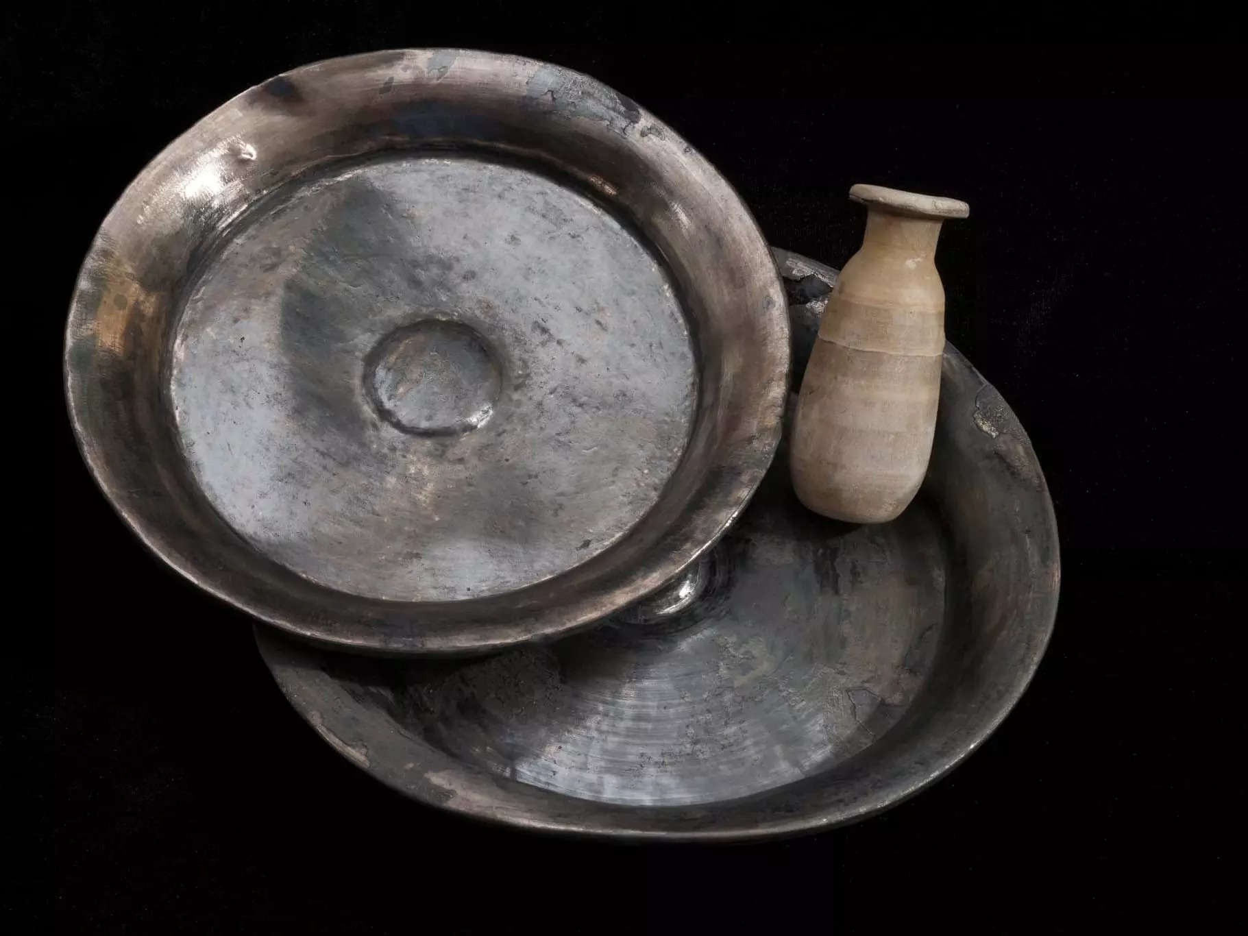 A picture shows two silver plates and a small alabaster container placed on a black background for display.