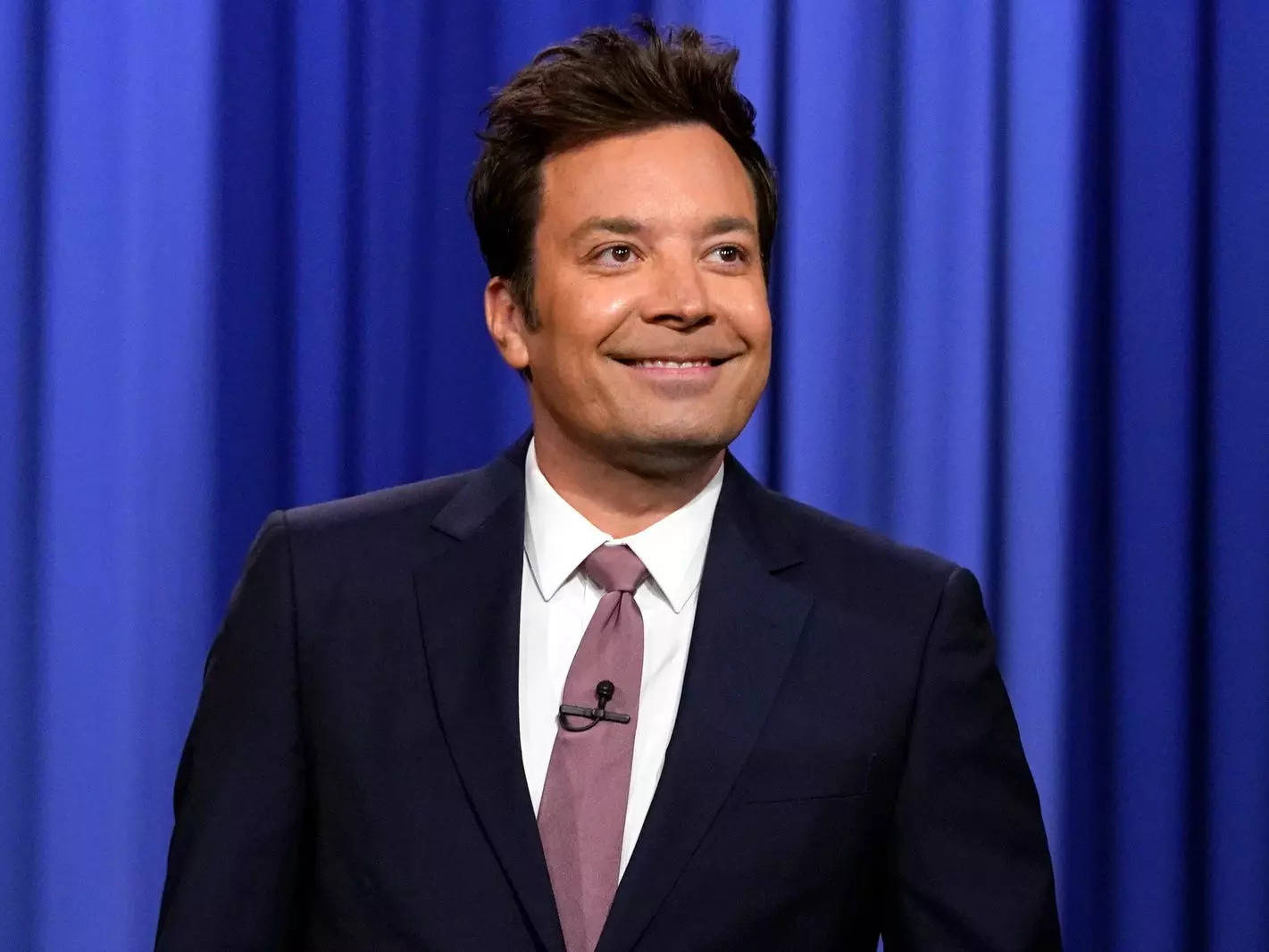 Jimmy Fallon in a suit and tie in front of a blue curtain.