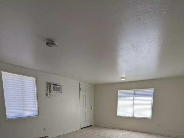 The living room has a wall air conditioning unit, two windows, and a tile floor in a neutral color.