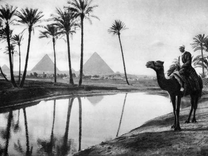 Circa 1920s: A camel rider basks by an oasis near Cairo, with the pyramids behind.