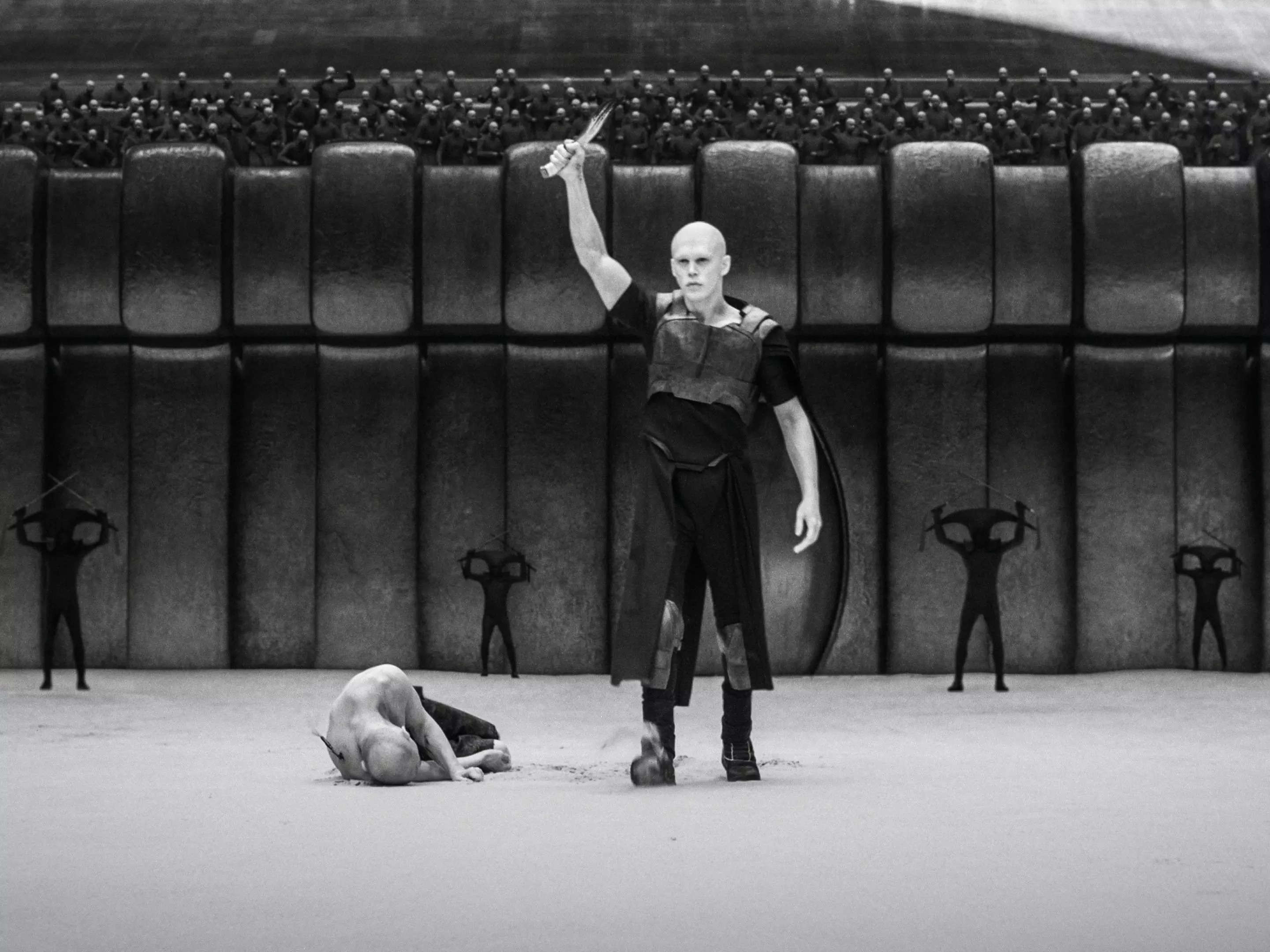 austin butler as feyd-rautha in an area, holding up a bloody knife while a man lies in the sand at his feet. the scenery, including the walls and crowd, are all in black and white