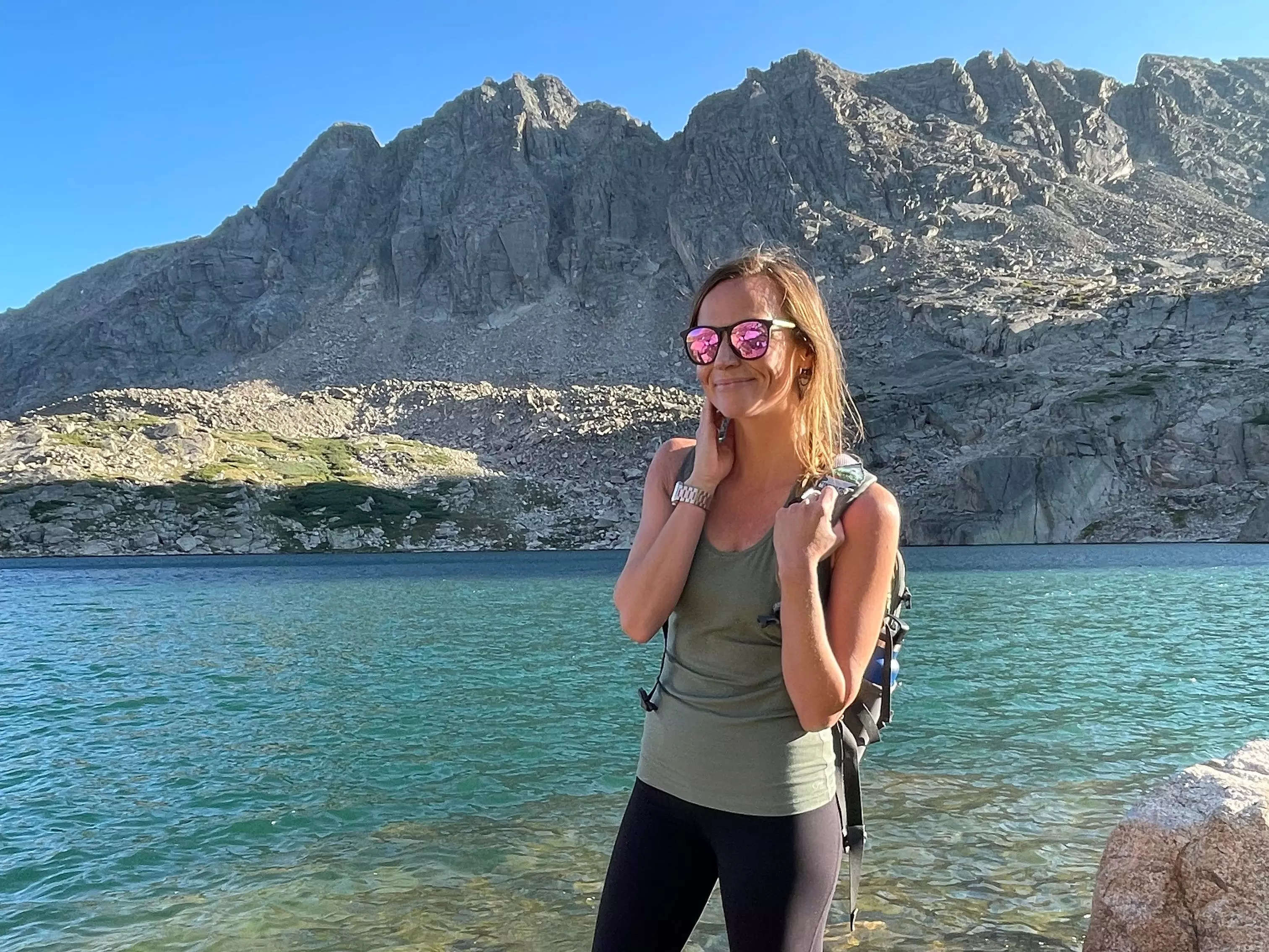 Emily, wearing sunglasses, a backpack, a green tank top, and black leggings, standing on a rock in blue water surrounded by mountains.
