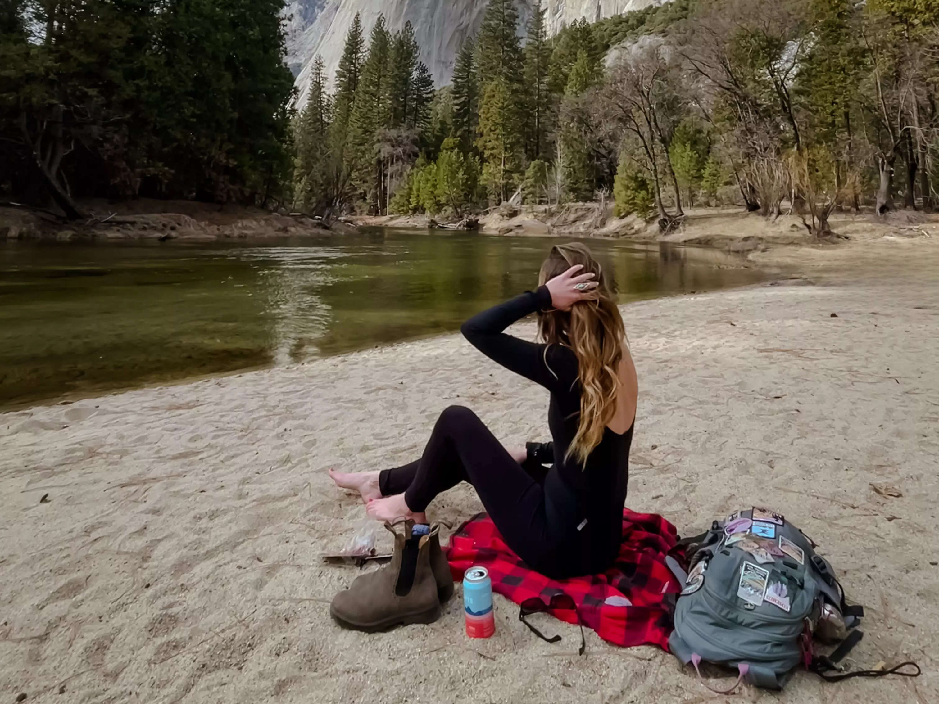 Emily sits on a black-and-red-checkered flannel looking out at water, trees, and mountains. Surrounding her are her sunglasses, boots, sunglasses, and a backpack covered with patches.