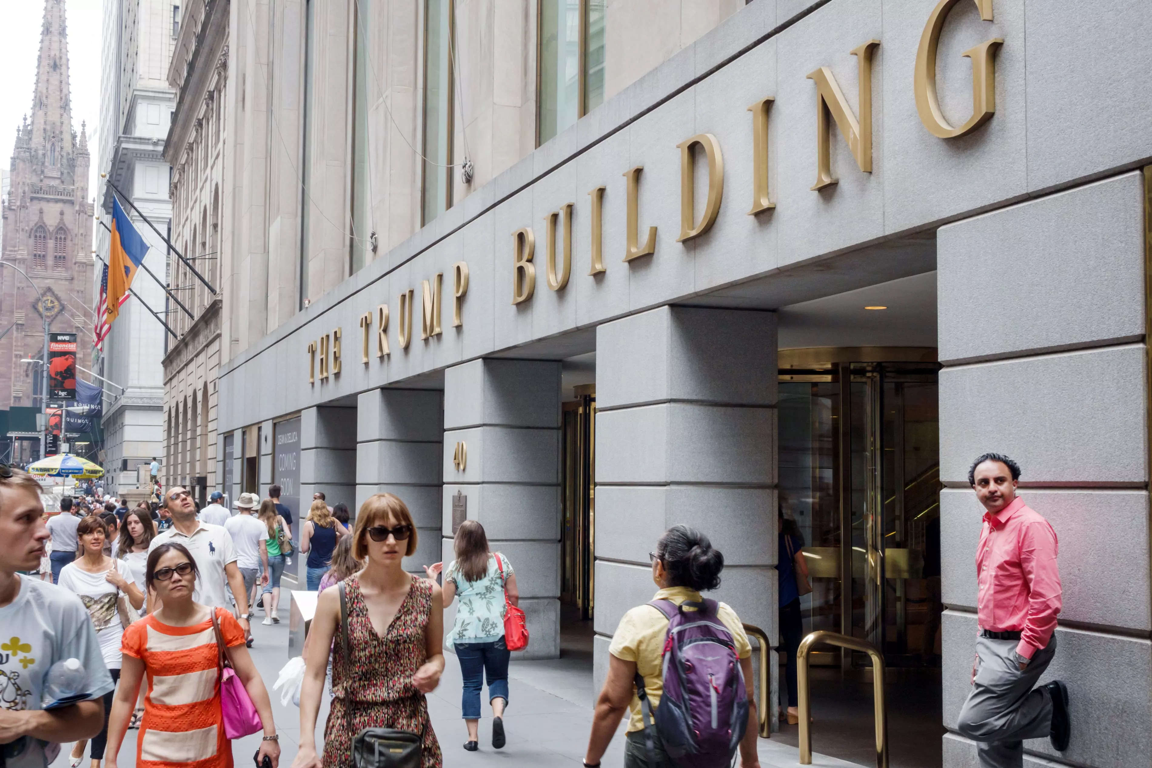 A crowd of New Yorkers walk past the gray facade of 40 Wall Street where "The Trump Building" is spelled out in gold letters