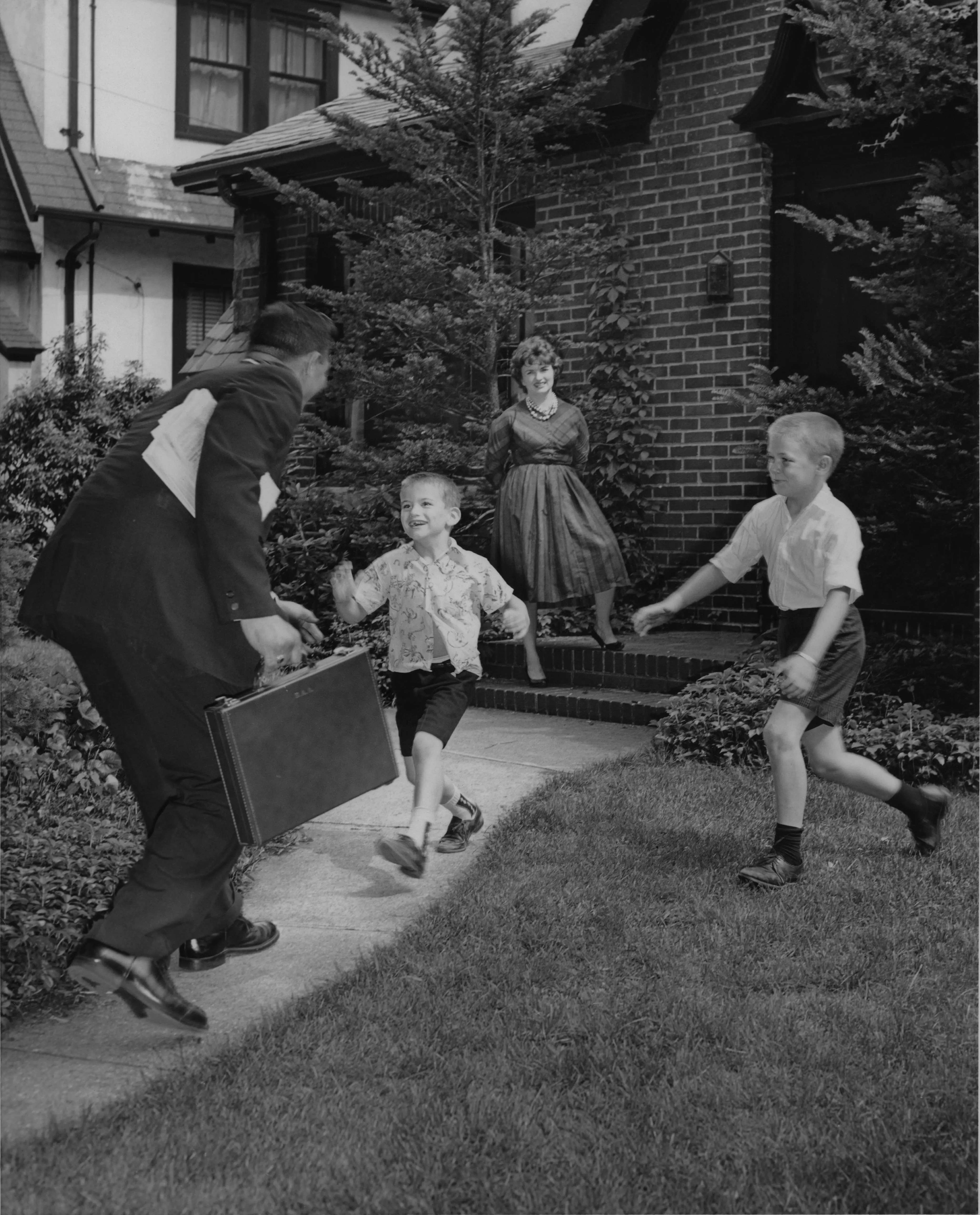 Two young boys in shorts run to greet Dad coming home from work while Mother is standing in the background. Circa 1960