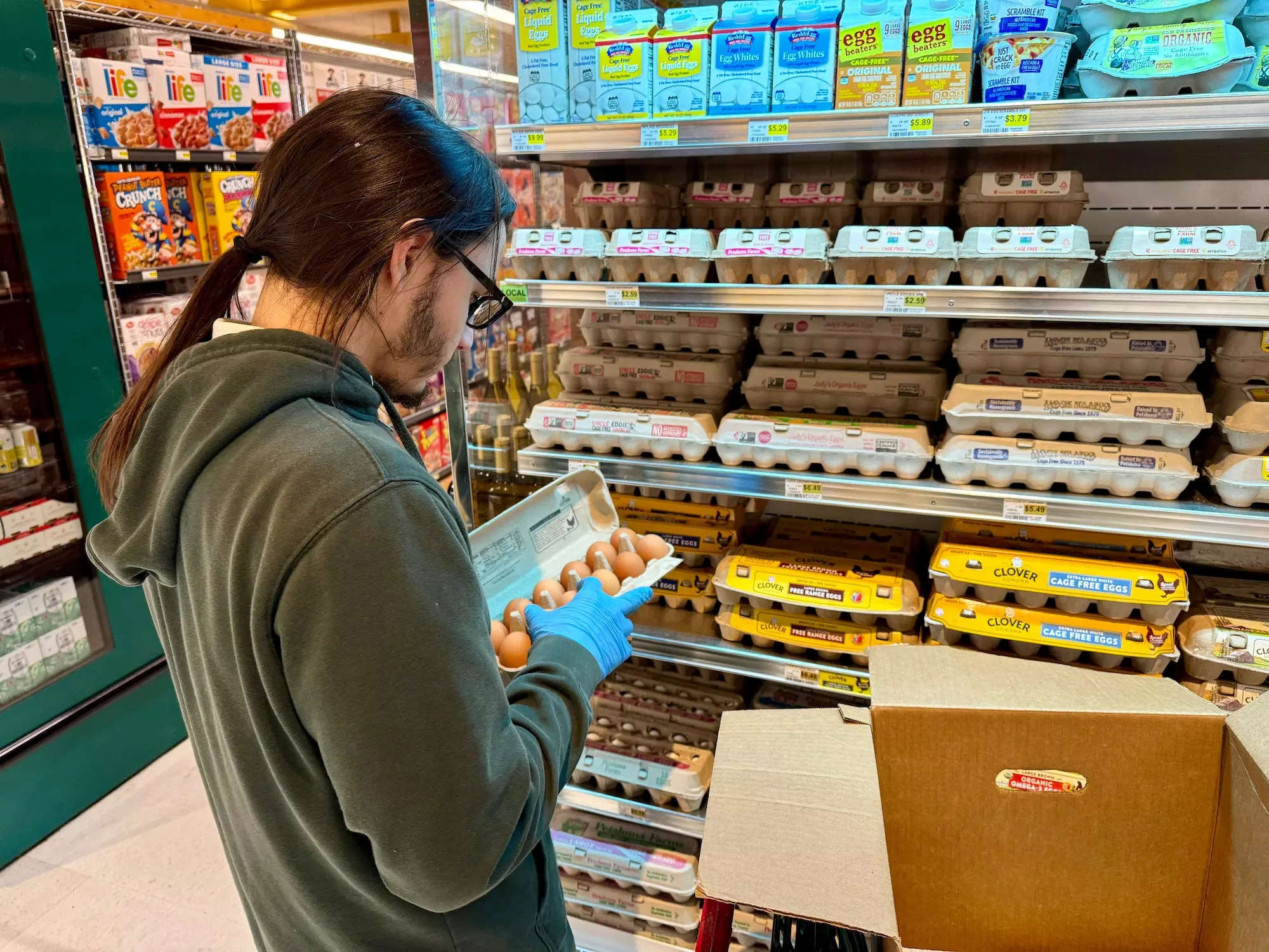person with ponytail green hoodie medical gloves inspects eggs in a grocery story