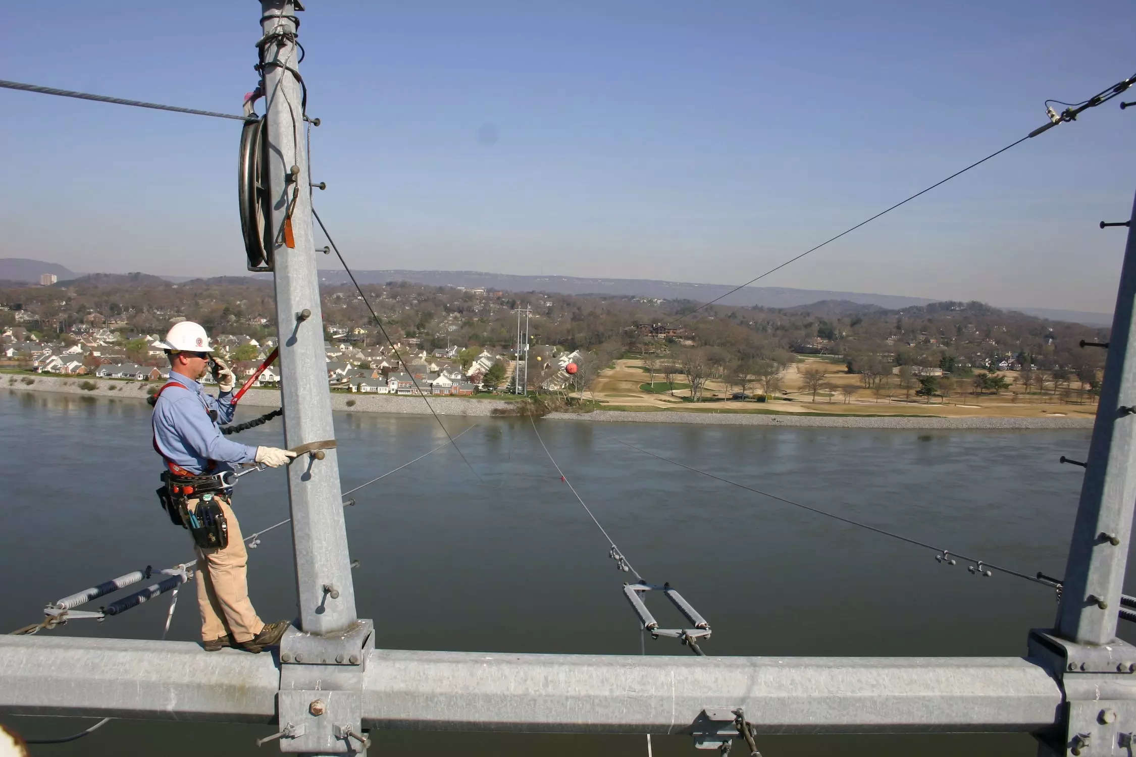 worker in hardhat wearing harness stands on large metal frame above a wide river with homes on the other side