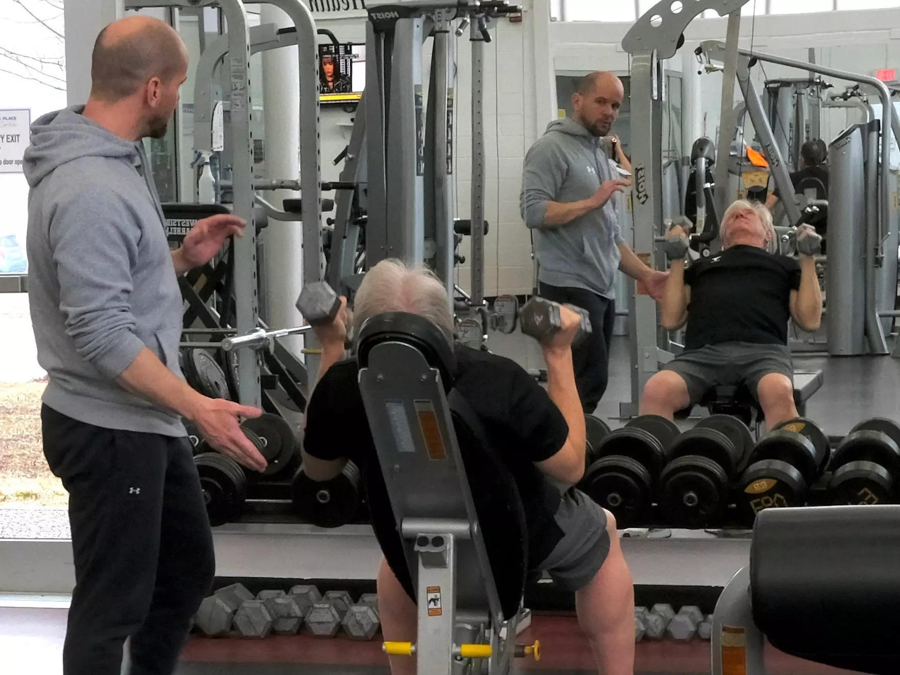 Barry sits on a workout bench and lifts weights. His trainer spots him and coaches him along.