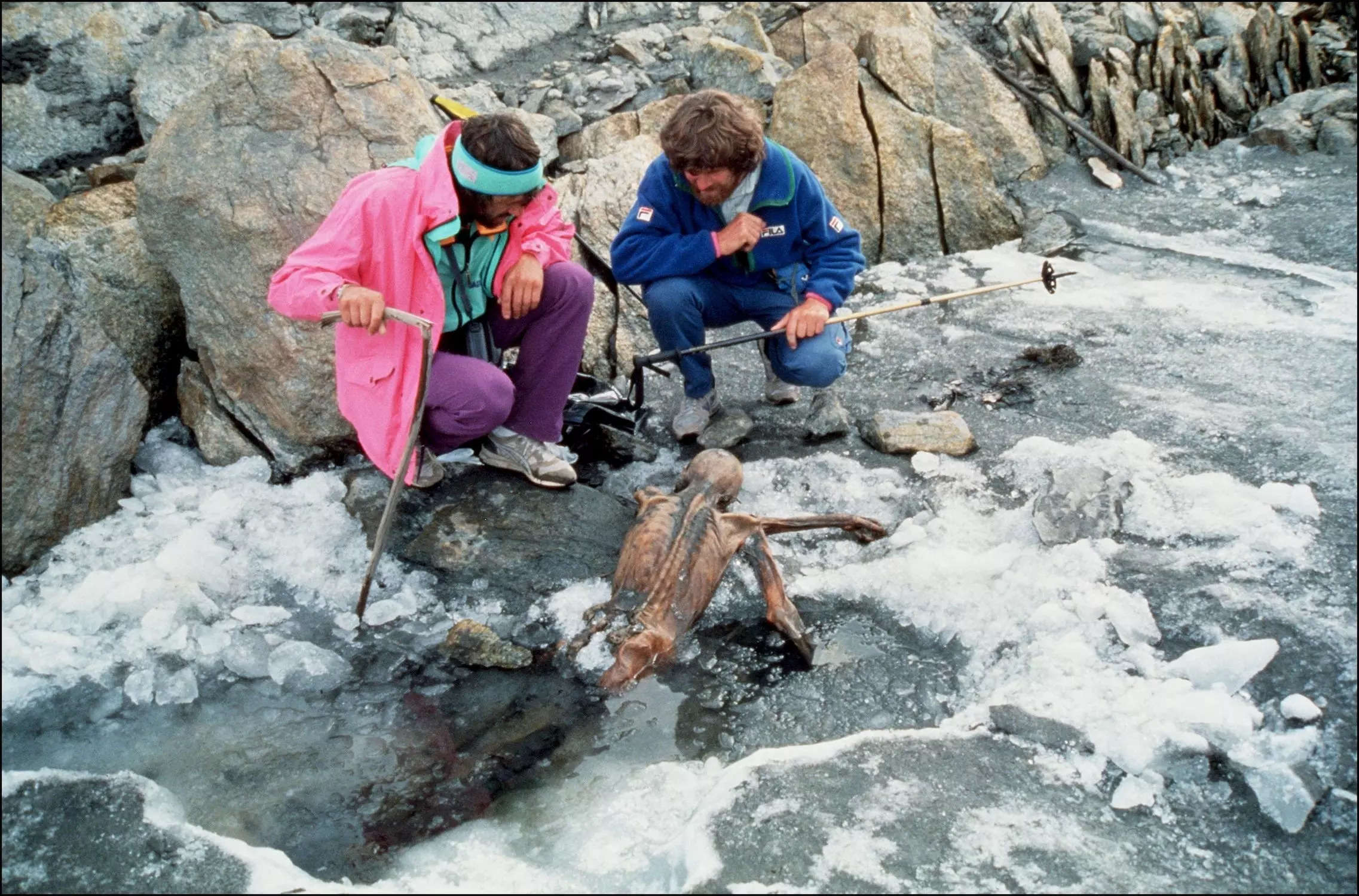 two men with shaggy hair in 90s hiking clothes crough on melty ice beside a facedown mummy positioned as if it