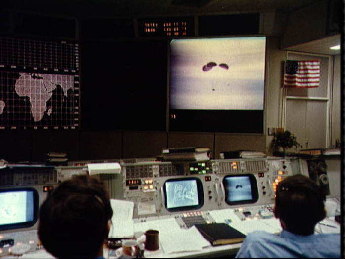 A large screen in front of the mission control room shows the spacecraft with its parachutes deployed.
