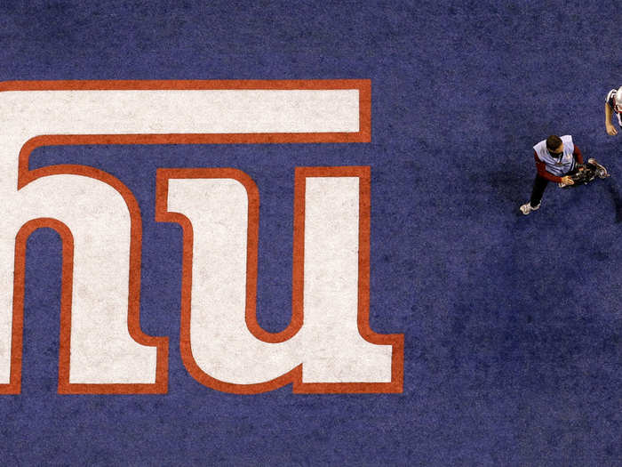 Tom Brady runs off the field past a Giants logo, with a cameraman in his face