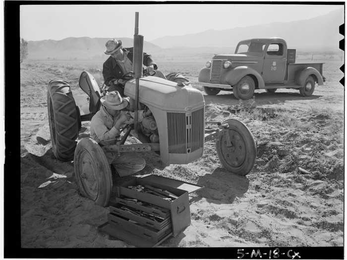 Here, a mechanic repairs a broken down tractor while the driver looks on.