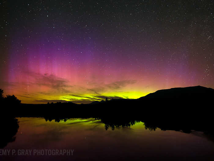 A colorful aurora illuminates the horizon while stars dot the night sky. A calm pond located west of Baxter State Park, reflects the night sky and the trees and hills, which separate Earth from sky in this image “Abol Bridge Aurora”.