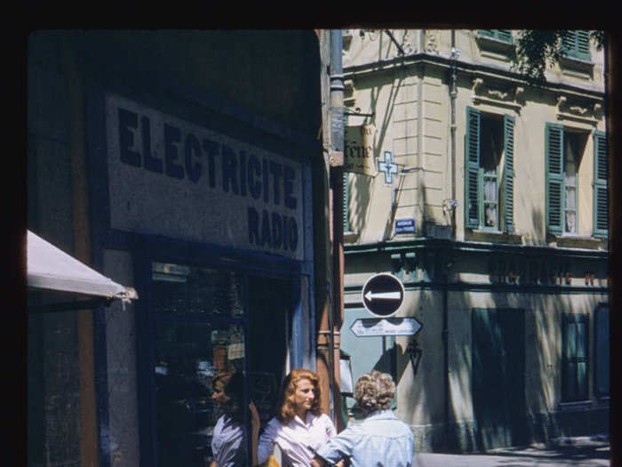 Two women dressed in elegant sixties attire stand outside an electric radio shop in Vence.