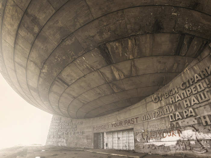 The outside of the building is covered in Bulgarian Cyrillic letters, many of which have fallen off. The stanzas say things like "On your feet, despised comrades, on your feet, you slaves of labor! Downtrodden and humiliated, Stand up against the enemy! Let us, without mercy, without forgiveness, yes, we take down the old, rotten system!"