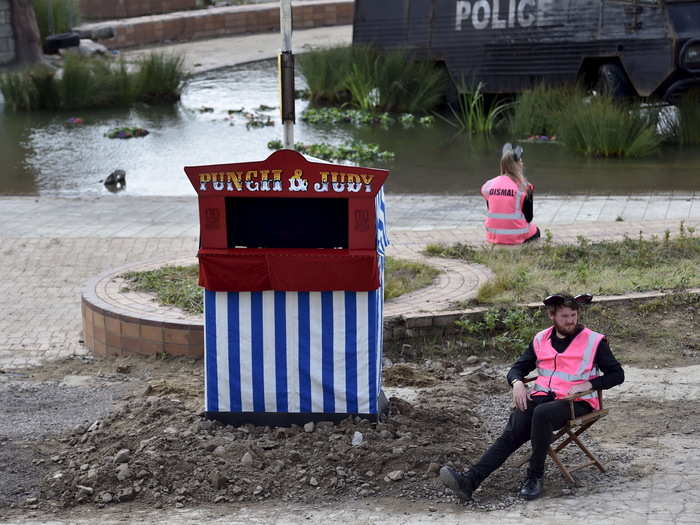 This bored attendant sits next to the "Punch and Judy" puppet show while an armored police vehicle rusts in the lagoon behind.