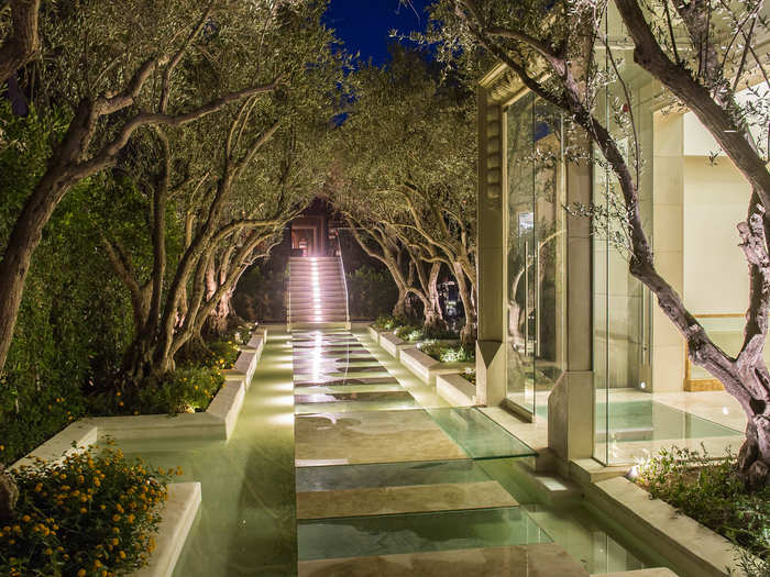 Guests enter through a floating glass-floor walkway lined with olive trees.
