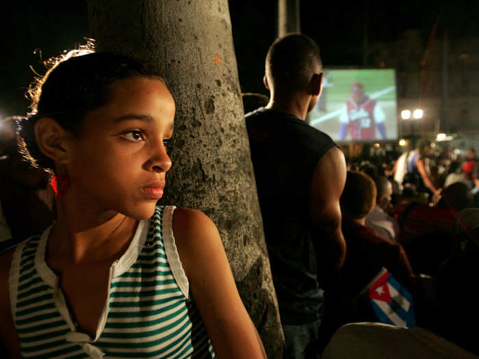 A girl leans against a tree in Havana while a group has gathered nearby to watch their country