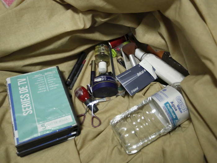 Nail polish, makeup, body lotion, a water bottle, and DVDs are seen on bed sheets of a bed inside the safe house.