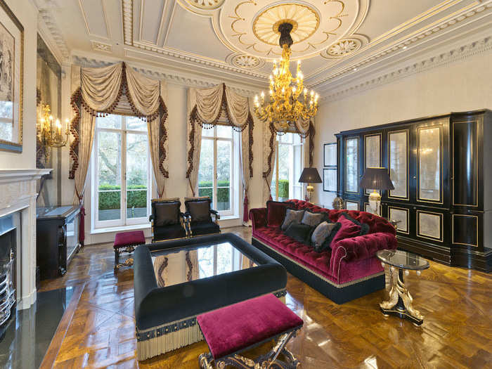 The living room oozes wealth, with a gold chandelier, plush furniture, and ornate ceiling decorations. Natural light pours through the large windows and door, which leads to a plant-lined balcony.