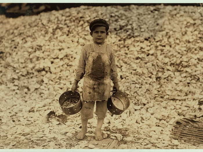 Manuel, the young shrimp-picker, 5 years old and a mountain of child-labor oyster shells behind him. He worked the year before. Understands not a word of English. Dunbar, Lopez, Dukate Company. Location: Biloxi, Mississippi, February 1911.