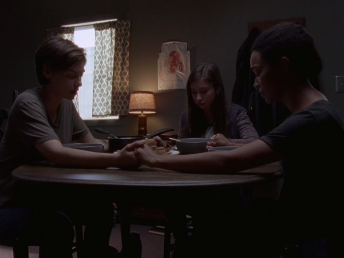 Maggie, Sasha, and Enid sit around a table holding hands to pray before eating a meal.