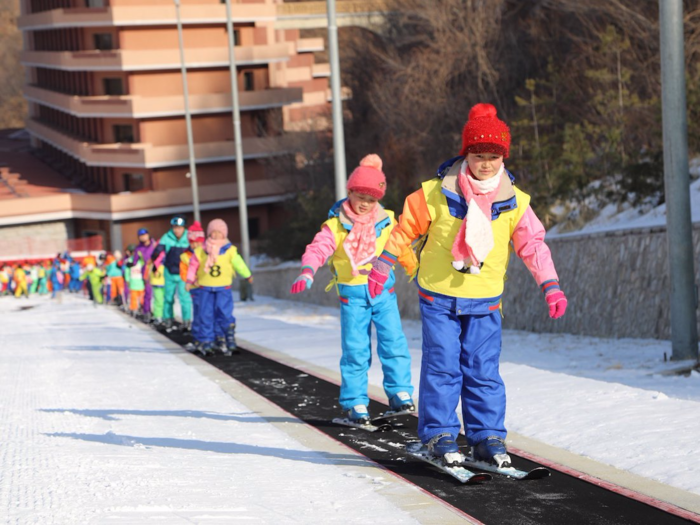 A large group of children taking part in a ski class ride a special convey-belt type track to climb part of the slope.