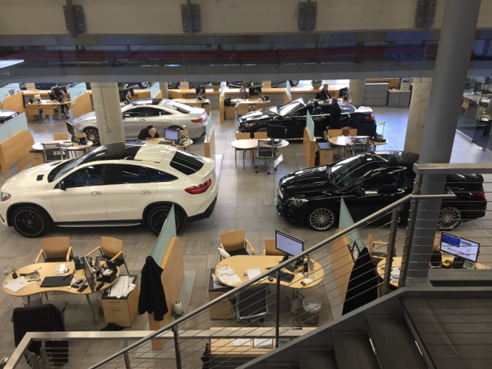 Open-air desks are arranged throughout the dealership and separated by rows of cars.