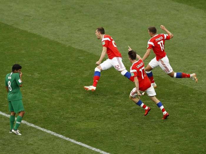 Russia celebrates a goal as a player for Saudi Arabia looks on.