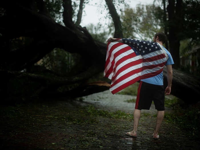 A Marine veteran named Josh stands with an American flag draped over his shoulders in front of a large fallen tree in downtown Wilmington, North Carolina.