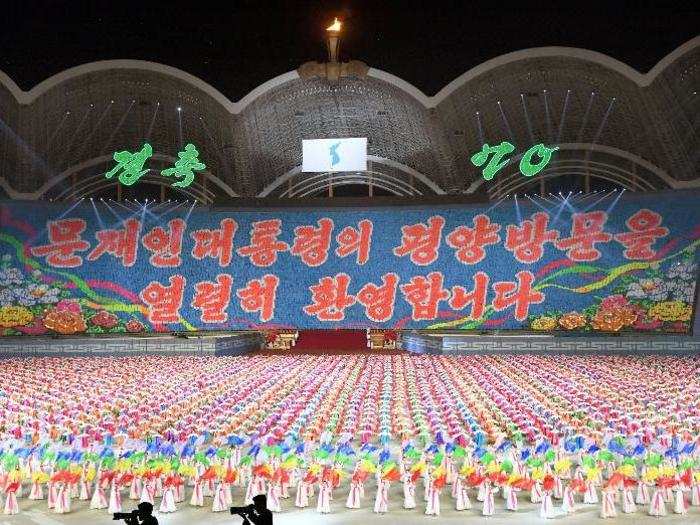 Thousands of performers, many of them children, hold up flip-cards to form a mosaic image that reads "We warmly welcome President Moon