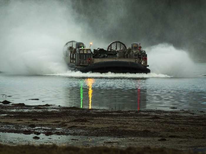 A landing craft carrying armored assault vehicles approaches the shore.