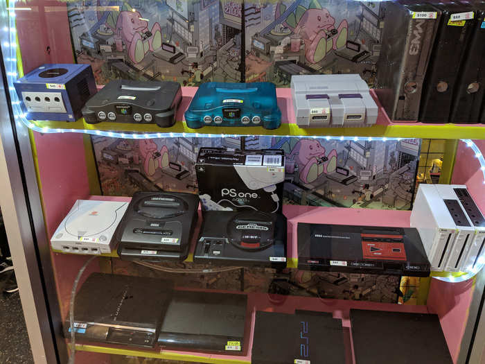 To the left of the plushies is an array of used game consoles, ranging from a Sega Master System to an Xbox One.