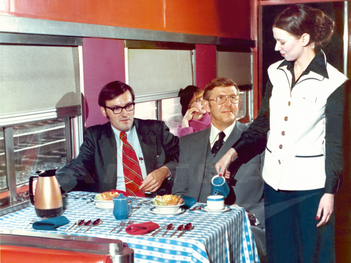 Around the mid-1970s, Amtrak redesigned the on-board service employee uniforms, which can be seen in this photo taken in 1976. By 1978, female attendants wore navy blue slacks and blouses or turtleneck pullovers, and white vests with blue trims, pictured below. The vests matched the jackets worn by men.