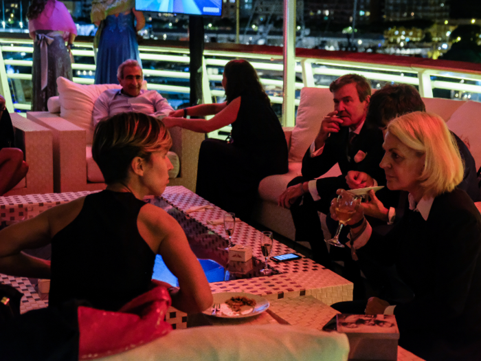 While most people remained downstairs, small groups had come up to relax on the upper deck of the yacht club, enjoying the relative quiet, the fresh air, and the views of the glittering port.