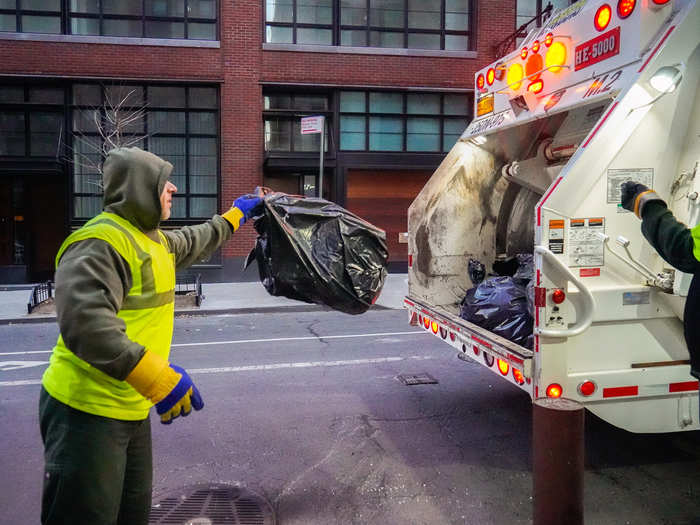 Sanitation workers must stand to the side of the hopper as they load trash into the vehicle.