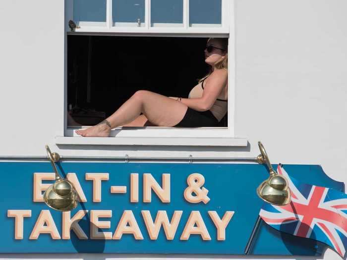 In a sea-side English town, a woman gets some fresh air and sunbathes from her window above a Fish and Chips restaurant.