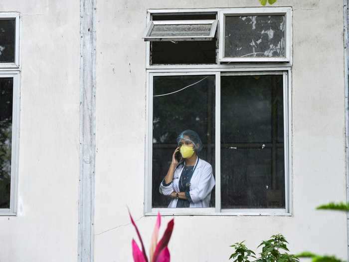 In India, a healthcare worker is photographed wearing a mask and uniform while talking on the phone in the window of a hospital.