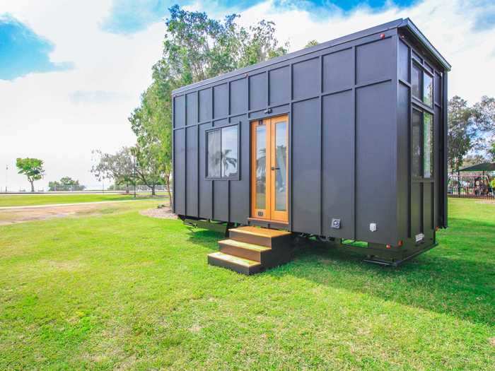 The tiny home has a dark exterior paint color that contrasts the bright interior filled with white walls and wooden floors, accents, and countertops.