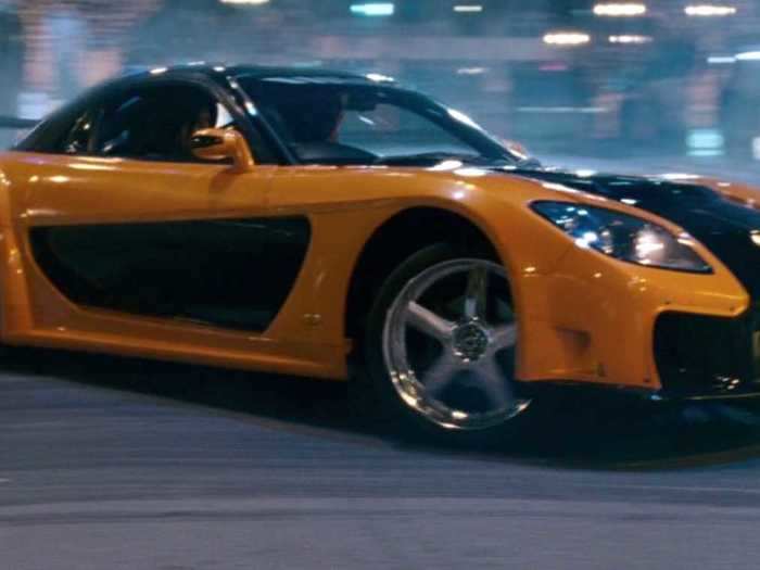 Han races through the streets of Tokyo in a black and orange Mazda RX-7 Veilside.